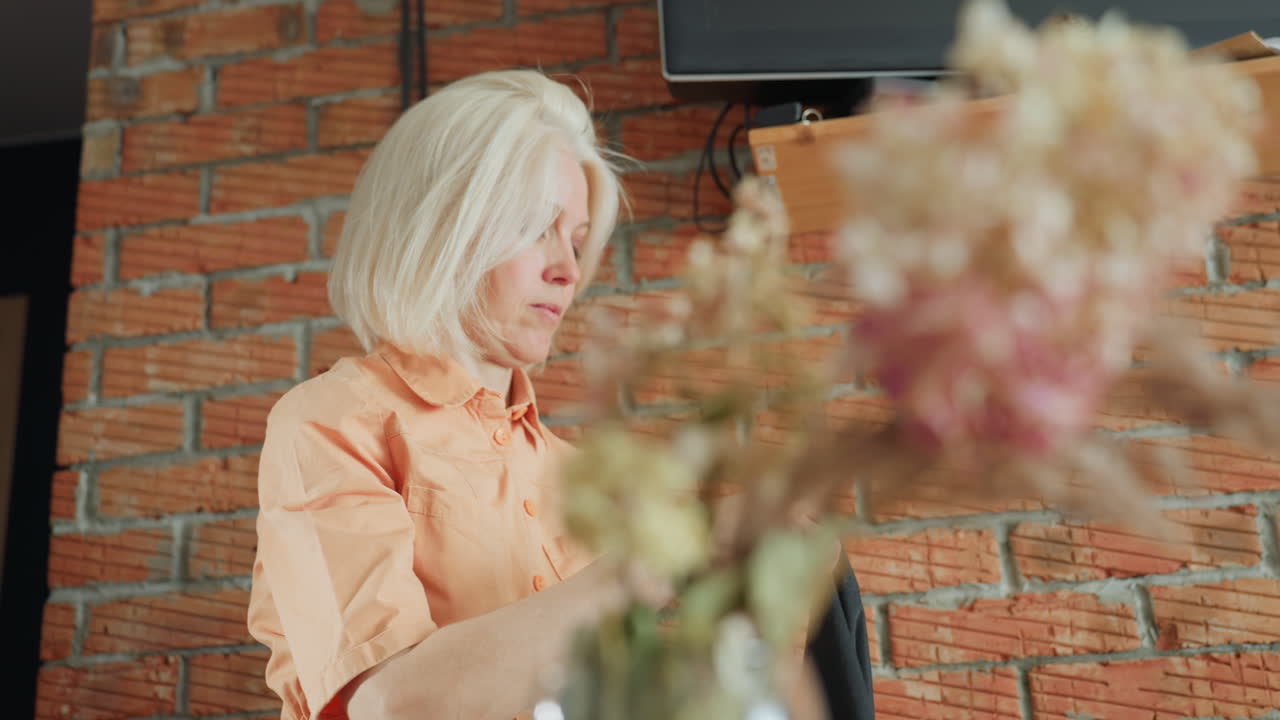 Decorator in orange shirt working with focus near rustic brick wall, holding black apron, blurred flowers in foreground creating artistic atmosphere, emphasizing preparation,dedication to design process