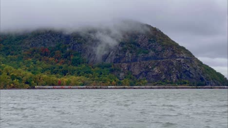 a freight train going along side a river during fall at the foot of a mountain as seen from the bank of the opposite side of the river