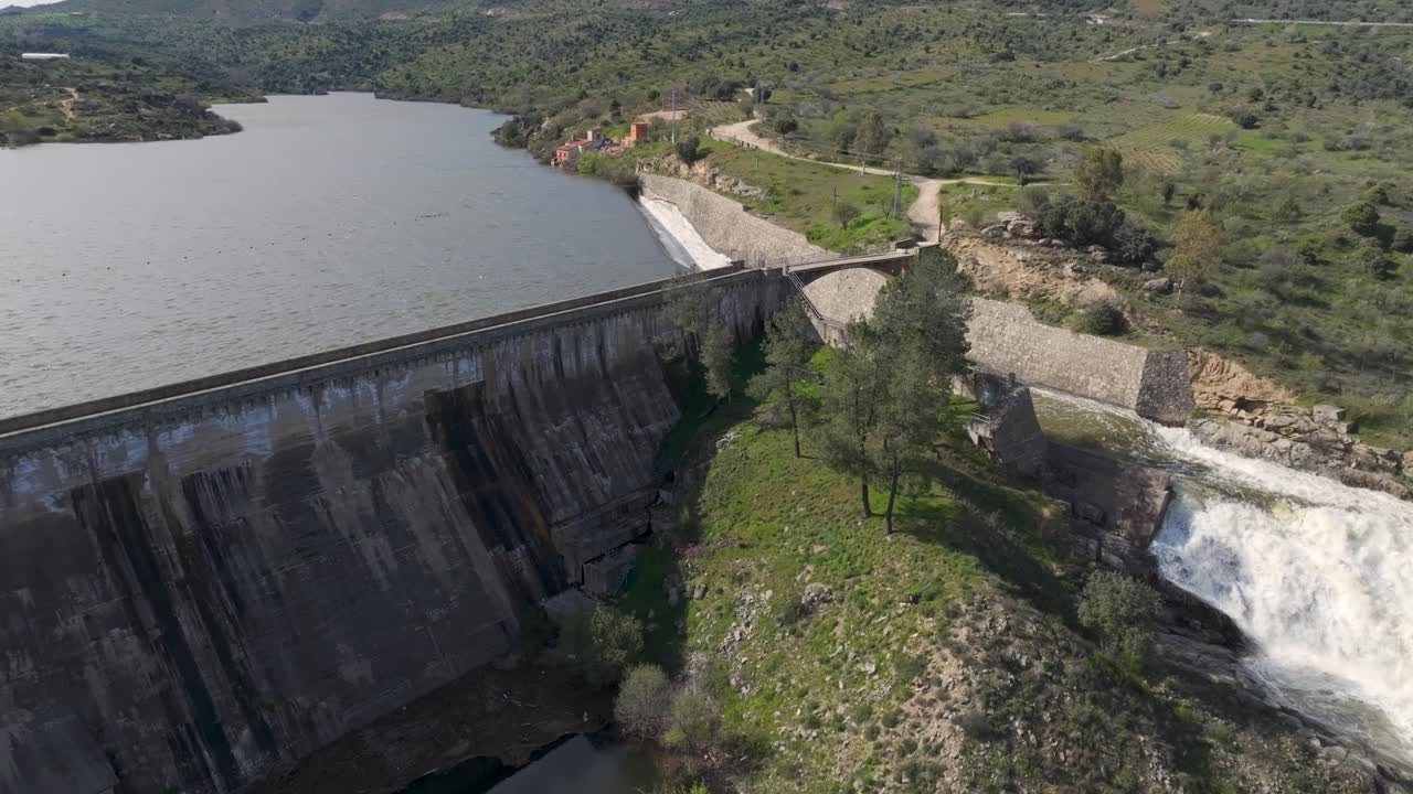 Ascending and reversing drone shot of a dam with lateral spillway releasing powerful water, forming a white horsetail-shaped waterfall and revealing the reservoir's size and scenic surroundings.