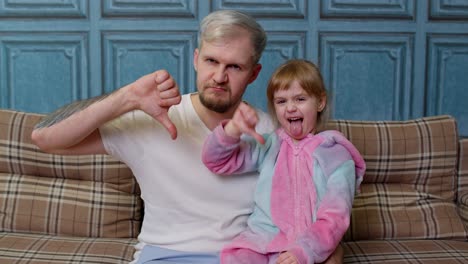 Displeased-father-and-child-daughter-kid-in-pajamas-sit-on-couch-in-room,-showing-thumb-down-dislike