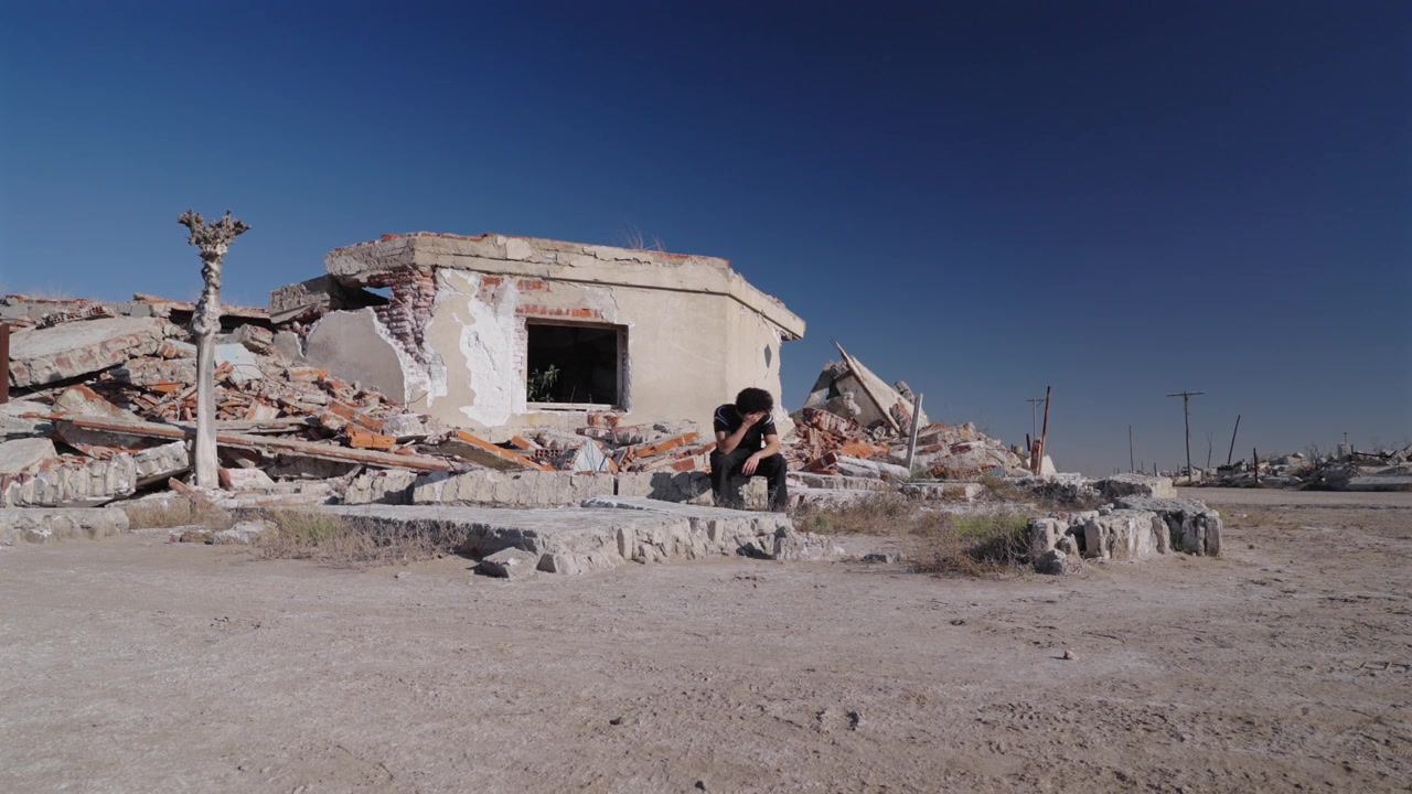 Inquadratura statica di un uomo triste e solo seduto tra le macerie e le rovine di un vecchio edificio a Epecuén, sotto un cielo pomeridiano di un blu intenso. Argentina.
