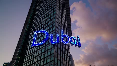 futuristic neon sign with dubai name hanging between skyscrapers at sunset with cloudy sky, modern architecture and illuminated buildings celebrating united arab emirates most important city