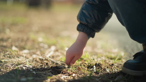 a young boy's hand, wearing a leather jacket, jeans, and boots, as he picks up a stick to dig into the soil, the stick breaks, and after trying again, he throws it away in anger