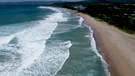 Olas-Rompiendo-En-Una-Playa-De-Arena-Con-Frondosos-árboles,-Luz-Del-Día,-Vista-Aérea