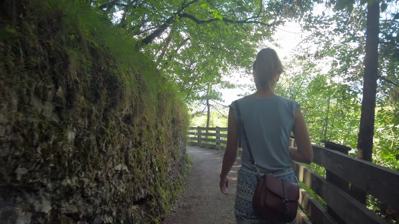Hiking woman is walking along a mountain path with a bag in summer. Following from the bak girl in blue t-shirt  walk In Deciduous Forest. Enjoying nature and Self-isolation.