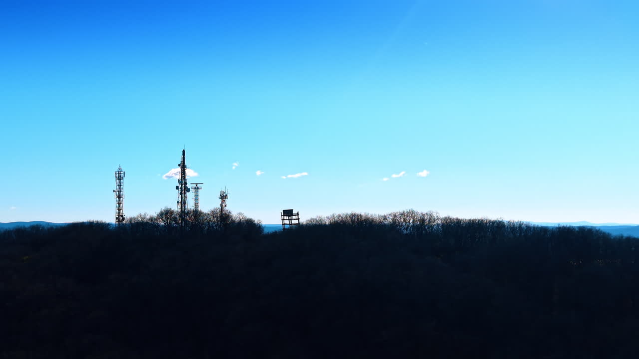 Silhouette di tralicci per telecomunicazioni su una collina al tramonto. Contorno scuro di torri e antenne di segnale su un crinale boscoso contro un cielo sereno