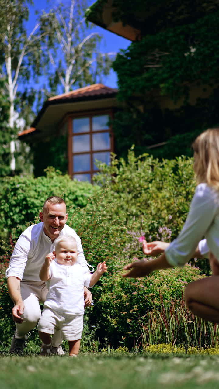 Lovely blond Caucasian child taking his first unsure steps by the grass in the garden. Supporting and loving parents are beside the child. Vertical video.