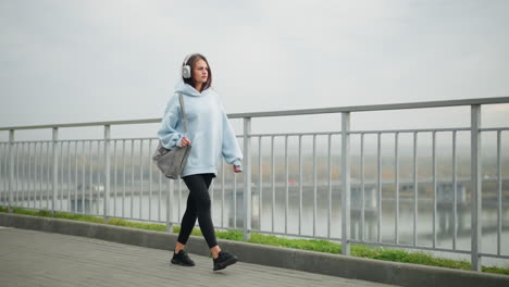 young fashionista in casual wear walking gently along street with iron rail, black shoes, bag, and headphones, background features bridge, flowing water, and green grass beside rail