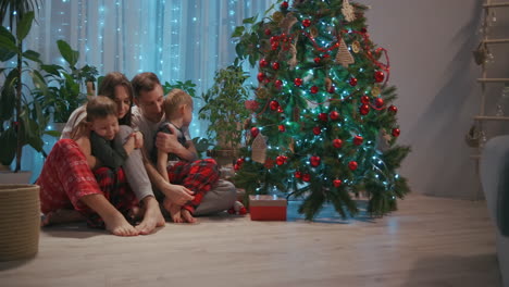 Una-Familia-Con-Dos-Niños-Se-Sientan-Juntos-Junto-Al-árbol-De-Navidad-Y-Se-Abrazan.-Amor-Y-Felicidad-Familiar-En-Nochebuena.-Padre-Y-Dos-Hijos-Se-Abrazan-Y-Miran-El-árbol-De-Navidad-En-Su-Sala-De-Estar.-Imágenes-4k-De-Alta-Calidad