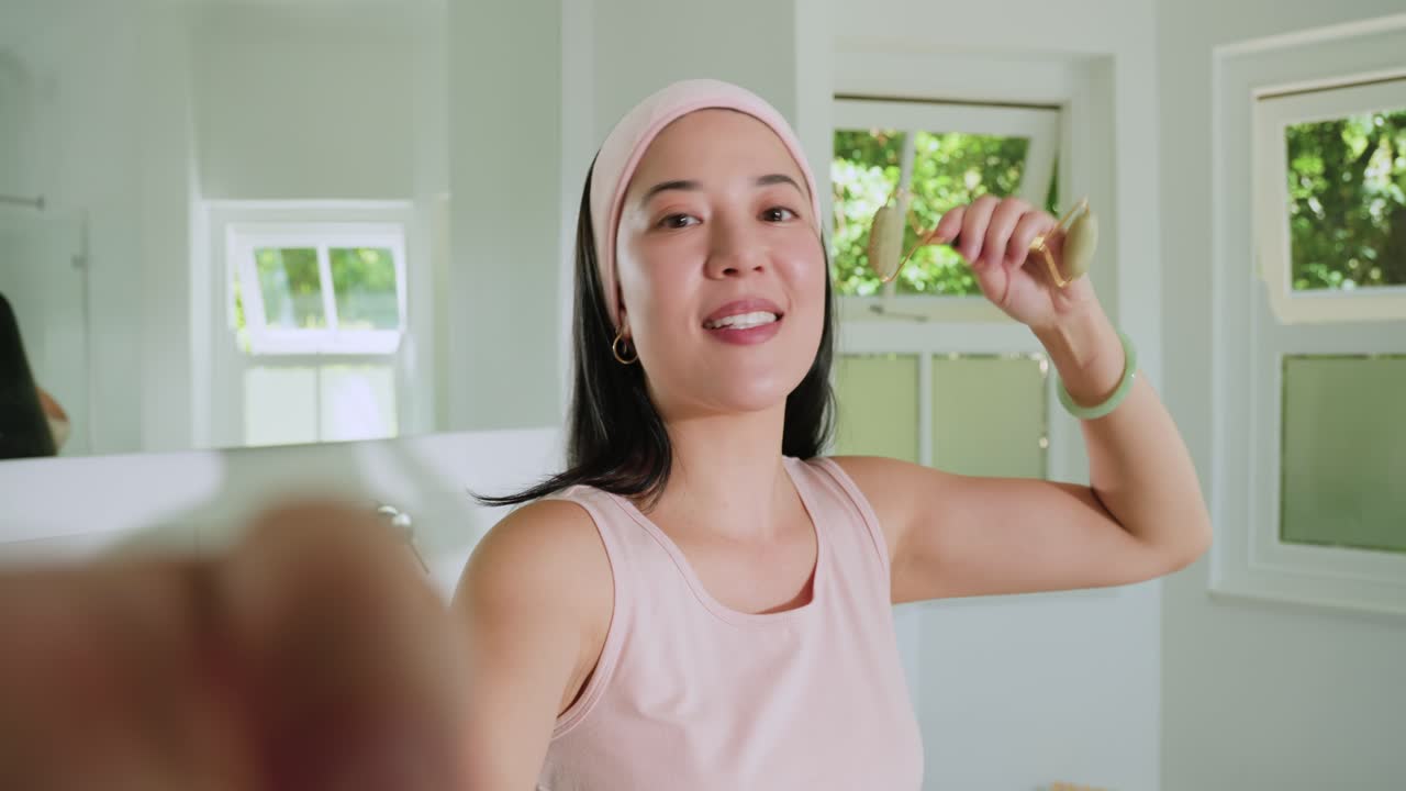 鏡の前でピンクのヘアバンドをつけたアジア人女性が、緑色のローラーを使って顎を転がし、スキンケアをしている。