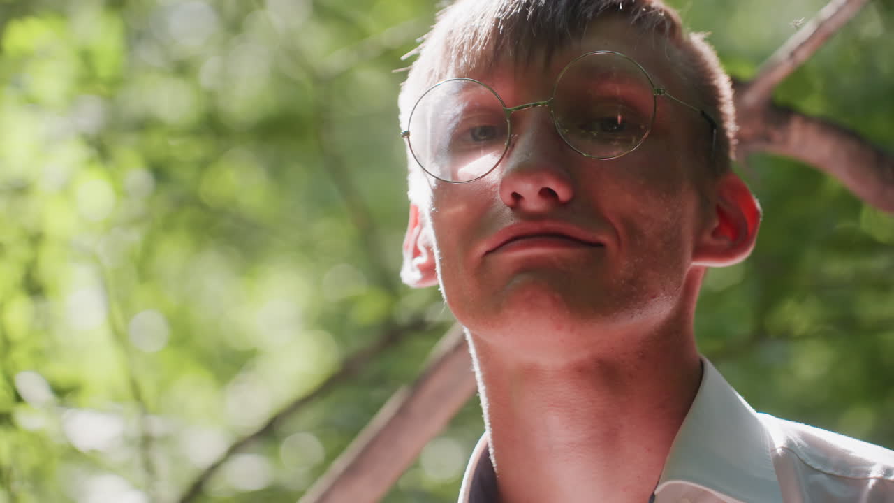 Portrait of man in white shirt and glasses standing in forest with focused gaze and light smile on his face, sunlight illuminating profile and highlighting peaceful expression