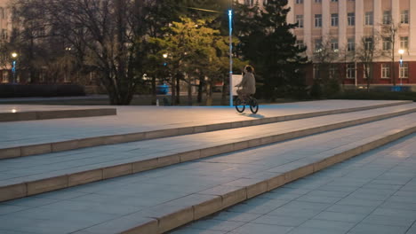 a person rides a bike up the steps of an urban park at dusk, he scene is illuminated by streetlights, with trees and buildings in the background