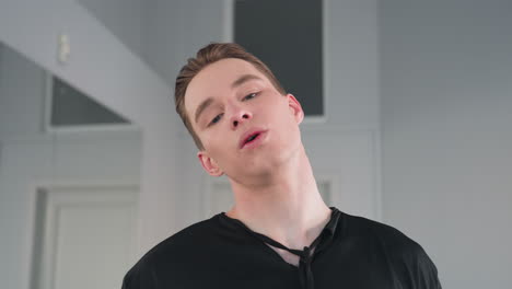 tired young man in black outfit standing indoors with eyes closed and arms folded taking deep breath in quiet moment of pause and relaxation inside minimalistic room with soft daylight reflections