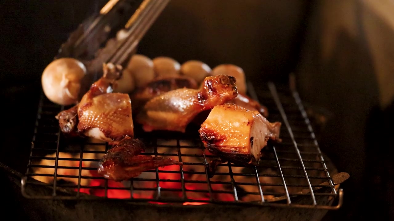 Close-up of chicken and sausages grilling over hot charcoal with metal tongs in Bangkok, Thailand.