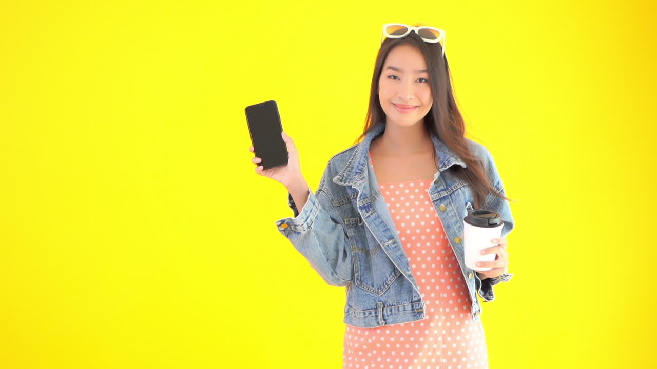 Chic Asian Female Showing Smartphone With Coffee To Go in Other Hand. Static Scene Isolated on Yellow Background For Chromakey