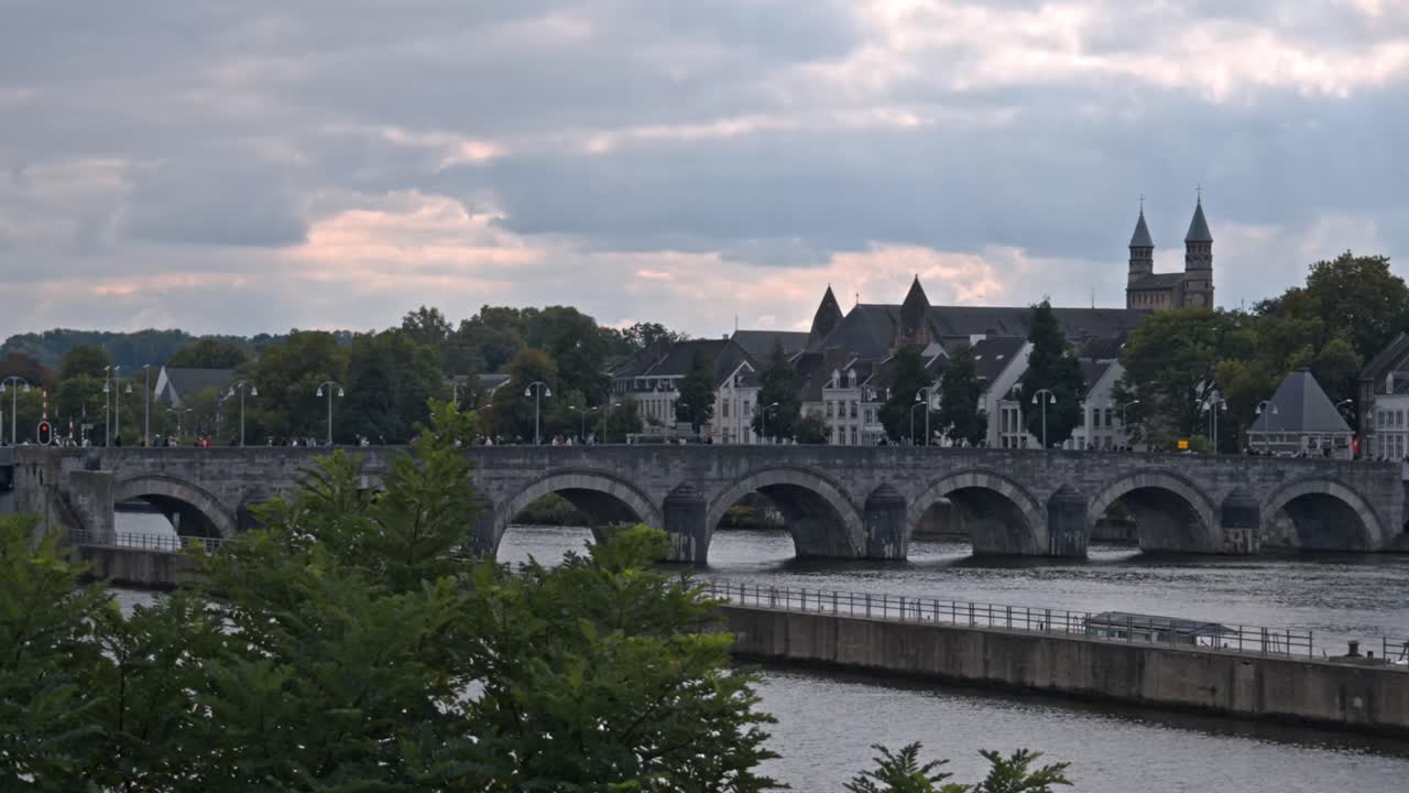 A wide shot of the Sint Servaasbrug (St. Servatius Bridge) spanning the Meuse River in Maastricht, Netherlands, under a dramatic, cloudy sky sunset