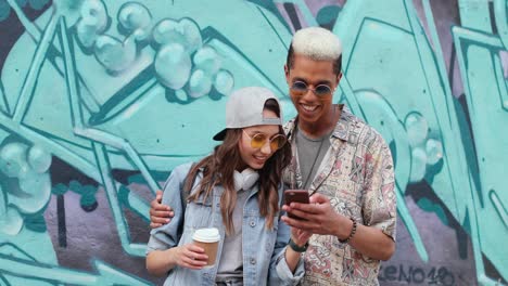 hipster couple in stylish clothes watching something on smartphone near a grafitti wall in the street