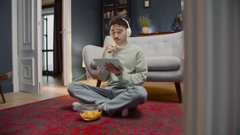 young man watching interesting movie on tablet while eating chips and sitting with crossed legs on the floor at home