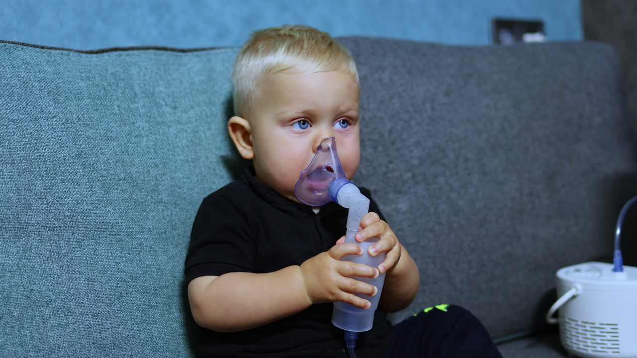 Küçük bir çocuk koltukta oturmuş inhaler ile nefes alıyor. Çocuklar için tedavi edici inhalasyon. Çocuklar için inhalasyon maskesi