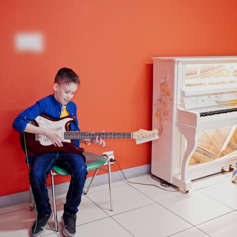 two happy boys are playing on musical instruments on the red wall background. one boy is playing on the acoustic guitar and another smaller boy is playing the piano.