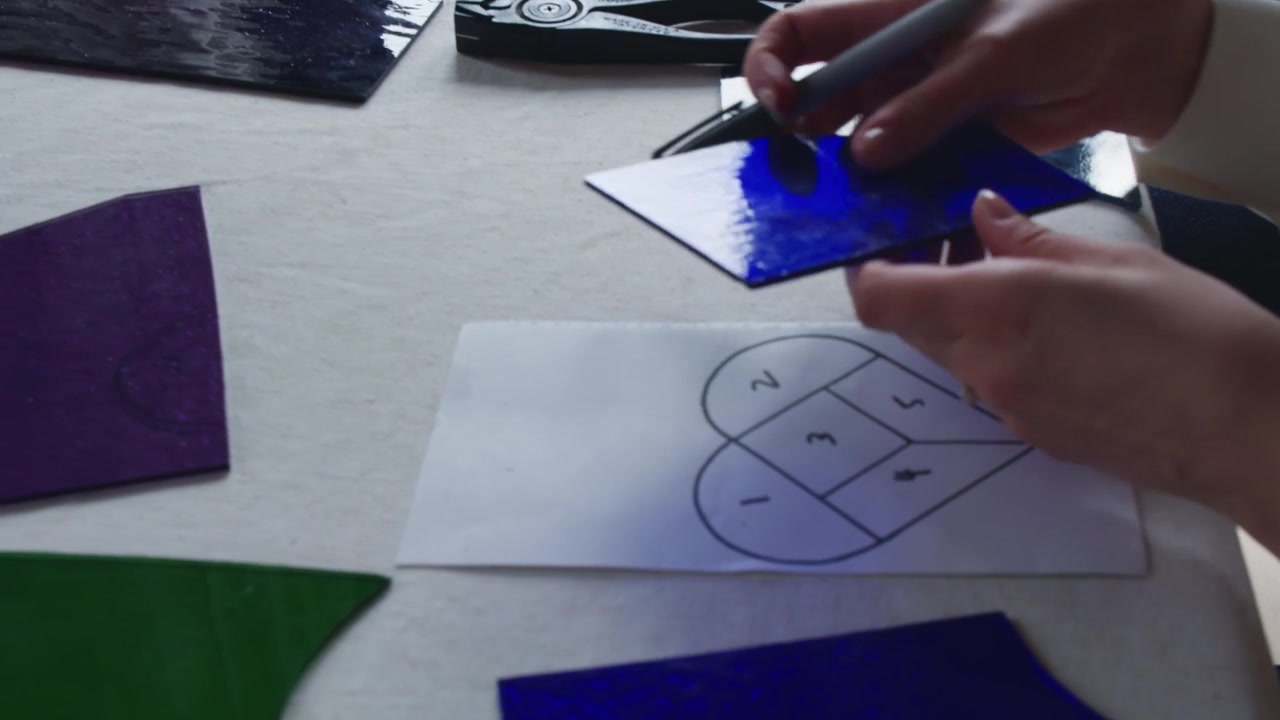 Two students work on a couples art project during a stained glass art class. The woman grabs a piece of blue glass and the camera follows her in for a close up of a heart template.