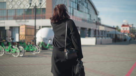 back view of woman in black coat and handbag walking on sunny urban street near green bicycles, with modern architecture and blurred background