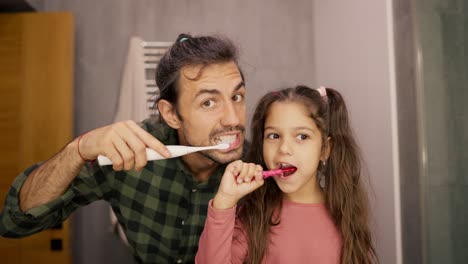 Portrait-of-a-brunette-man-in-a-green-checkered-shirt-brushing-his-teeth-with-his-little-daughter-brunette-girl-in-a-pink-dress.-They-look-in-the-mirror,-look-at-the-camera-and-brush-their-teeth-in-a-modern-bathroom