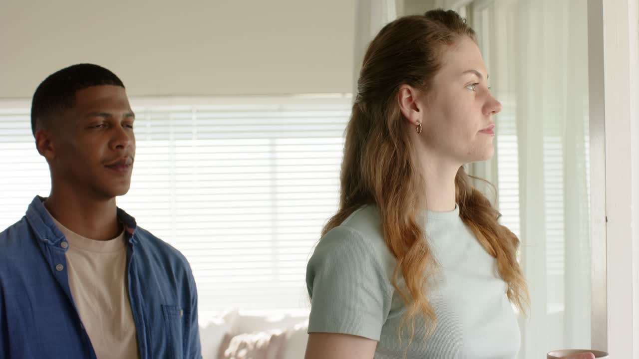 Diverse couple standing at home by window woman holding mug man entering both gazing sharing calm