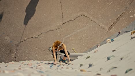 shot from above, a blonde girl in sunglasses, a black sports summer uniform and in special equipment for rock climbing and belaying climbs up a white climbing wall with multi-colored ledges and trains her rock climbing skills in the summer