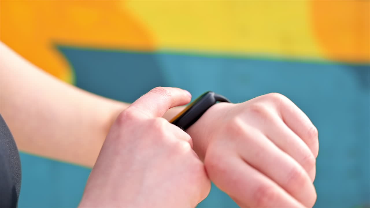Young woman at outdoors training in sportswear is on her fitness bracelet, earphones, multicolored background. Slow motion