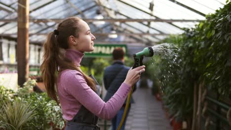 Young-attractive-female-gardener-in-uniform-watering-plants-with-garden-hose-in-greenhouse.-Slowmotion-shot
