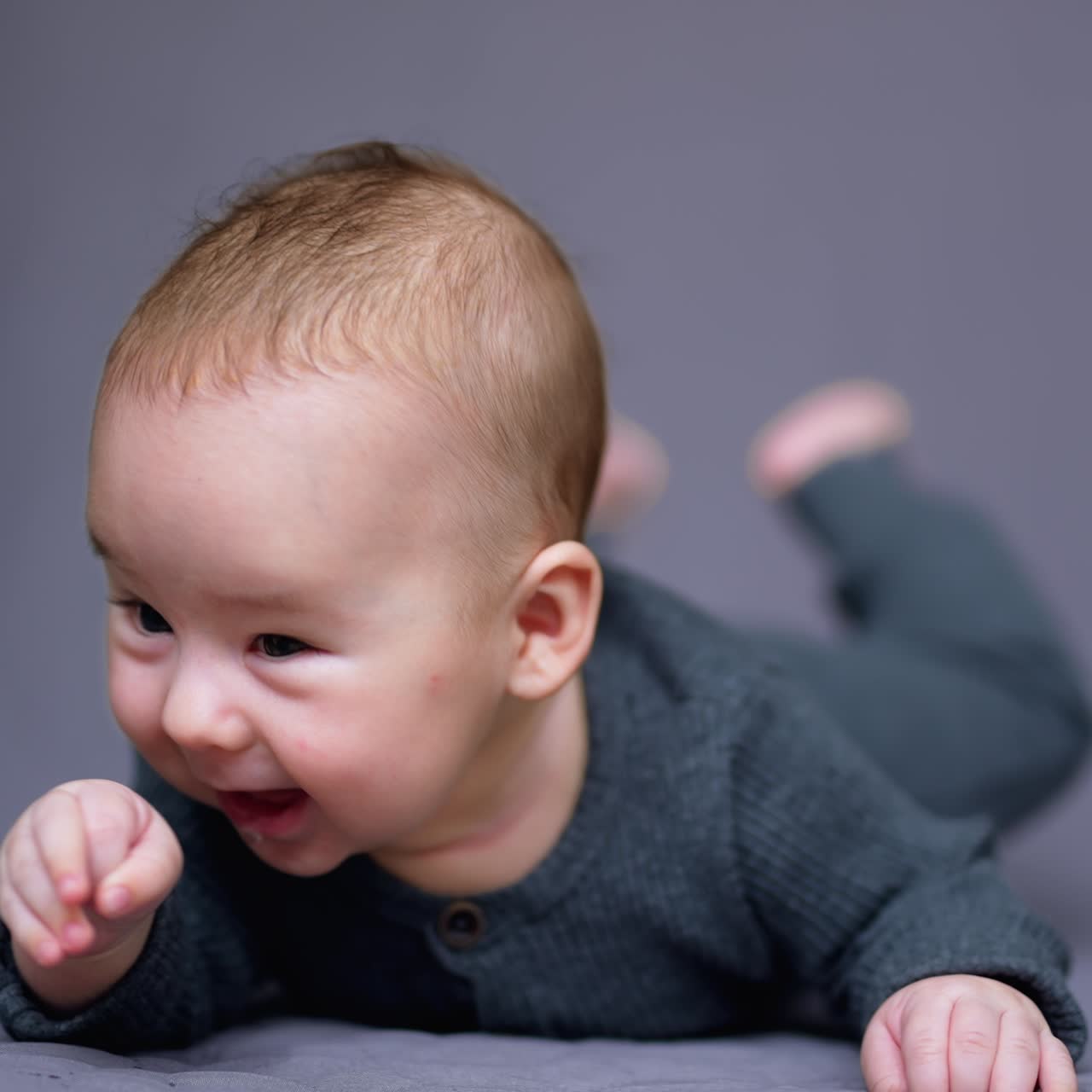 Adorable baby boy lies on bed and looks into camera. Beautiful child smiling sweetly. Grey backdrop