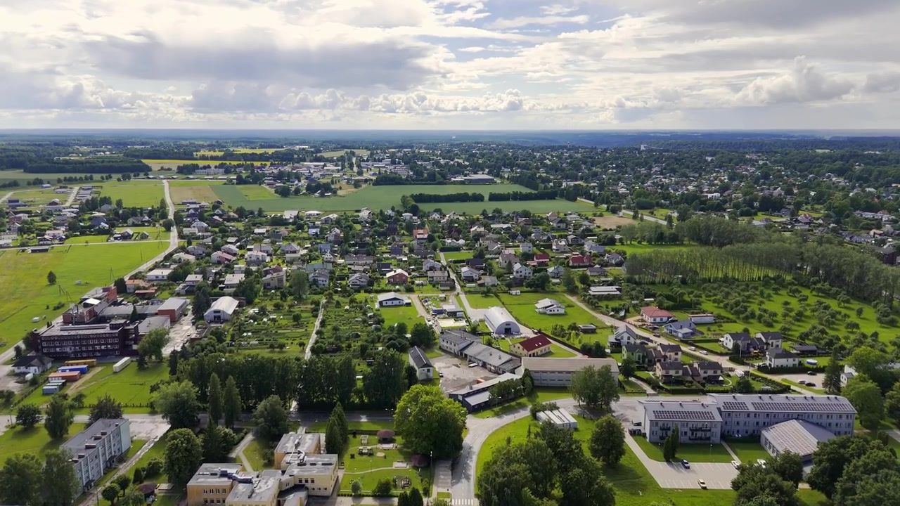 Vista aerea con drone su Sigulda, Lettonia, che mostra quartieri residenziali, campi verdi, strade alberate, edifici pubblici e un paesaggio urbano distante sotto nuvole estive.