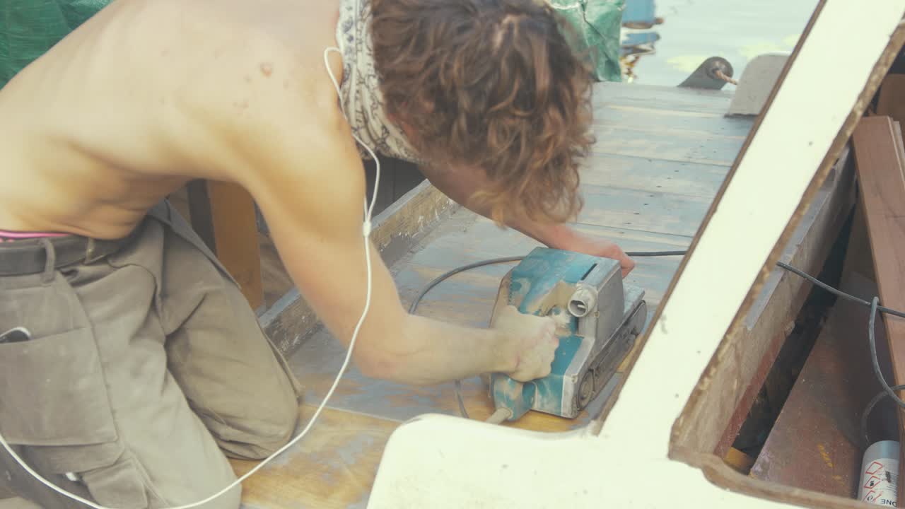 l'uomo leviga il fasciame del ponte in teak con levigatrice a nastro su una barca di legno in topless sotto il sole estivo