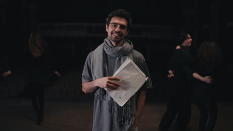 Portrait-of-a-happy-young-male-stage-director-with-papers-in-his-hands-with-a-scarf-and-T-shirt-on-stage-in-the-theater-during-a-rehearsal