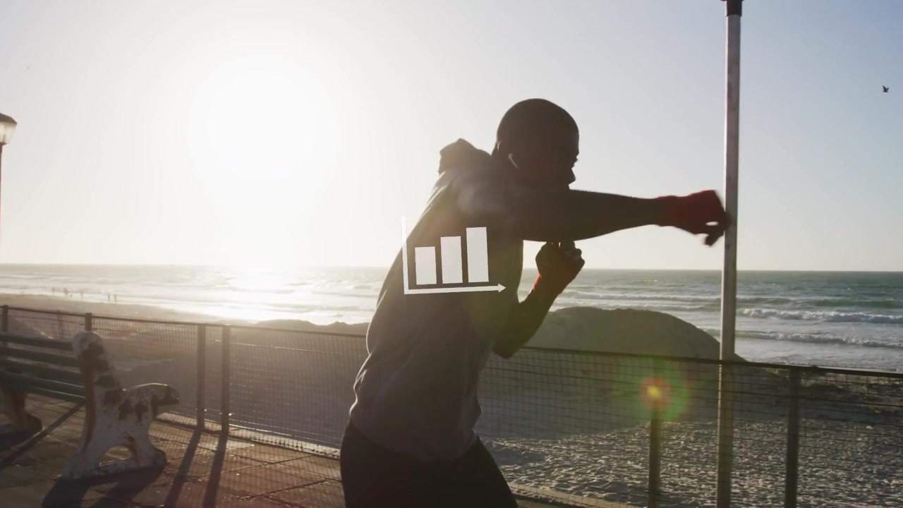 Vídeo vertical: Homem praticando boxe de sombra, vestindo moletom com capuz e calças escuras, treinando perto de um corrimão à beira-mar, gráfico.