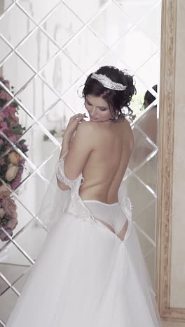 slow motion wonderful bride with naked back stands near flower arch in room with huge segmented mirror