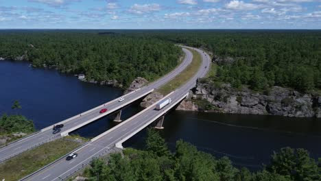 Toma-Aérea-Ascendente-Del-Puente-De-La-Autopista-Curva-Sobre-El-Lago-A-Través-De-Rocas-Y-árboles-Cerca-De-Gravenhurst-Ontario