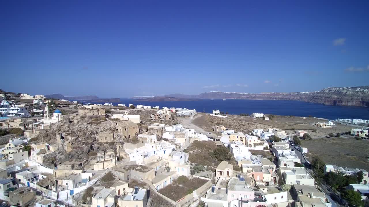 A picturesque Greek coastal village with white and stone buildings, overlooking the deep blue Aegean Sea and surrounded by rocky hills under a bright blue sky.
