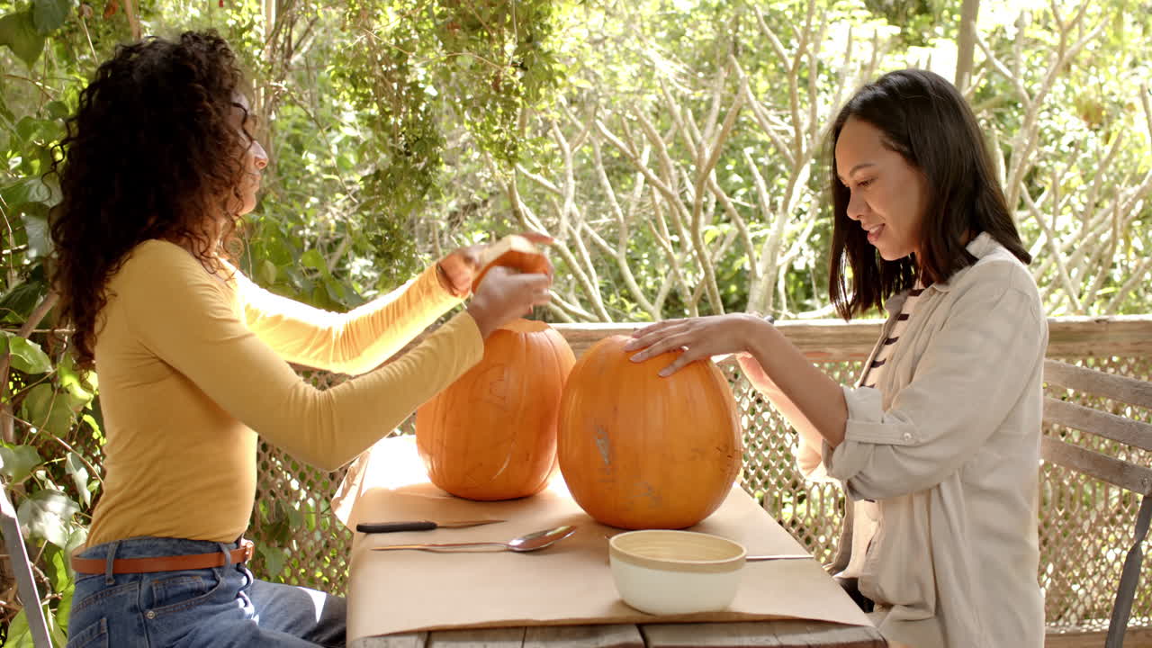 Halloween time, two multiracial female friends carving pumpkins on a porch