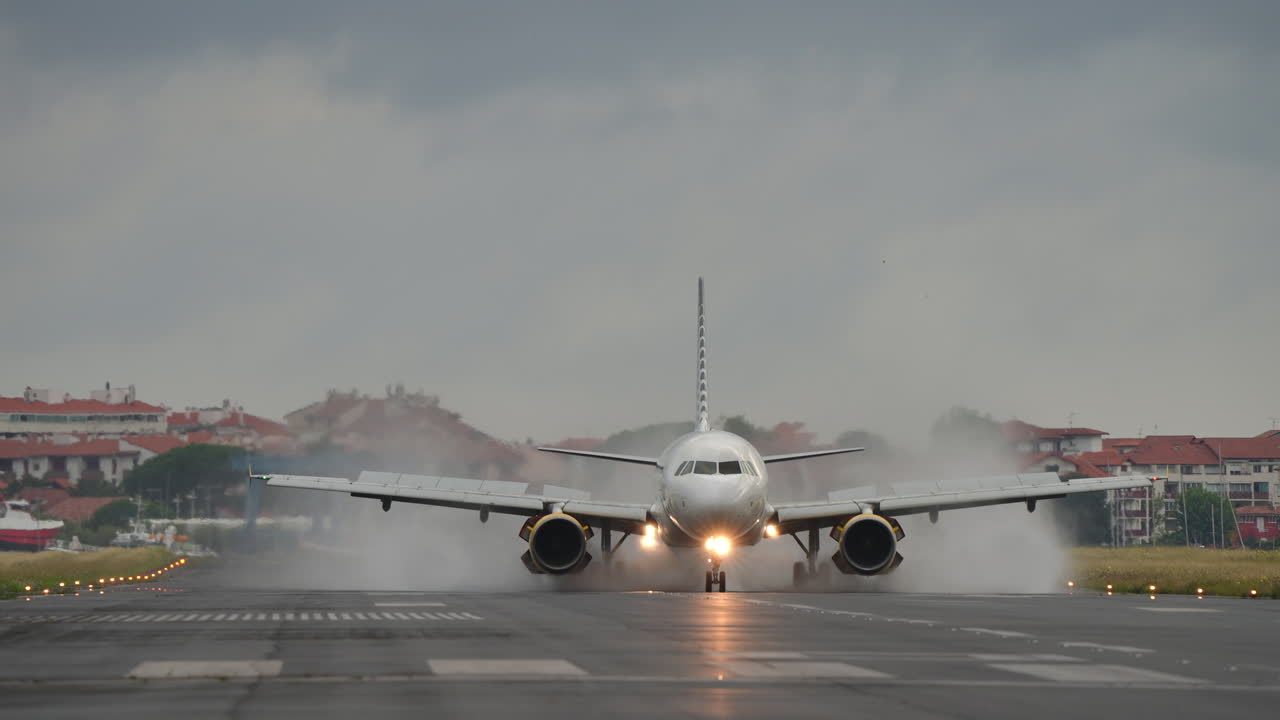 Airplane approaches runway, creating water spray on landing, with cloudy sky and urban background enhancing the scene