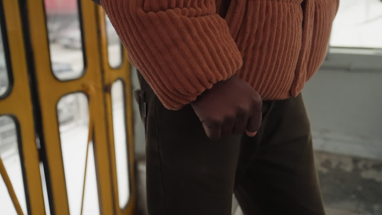 Primo piano della mano di un pendolare all'interno di un tram giallo, maglione di lana e pantaloni di velluto a coste, passeggero solitario che si tiene alla porta, tessuto testurizzato contro telaio metallico, luce invernale che proietta un'ombra morbida, umore da pendolare in attesa.