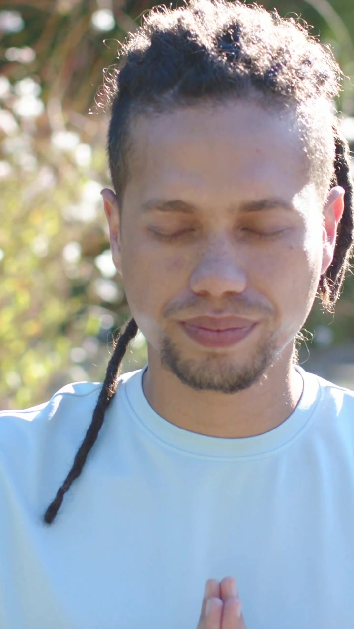 Video verticale: Con i palmi uniti, uomo che medita che respira in giardino, con maglietta blu e collana