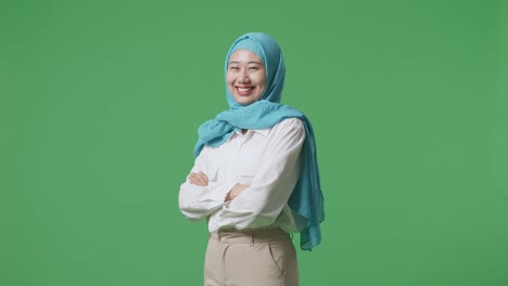 vista lateral de una mujer musulmana asiática sonriendo y cruzando los brazos mientras está de pie en el estudio de fondo de la pantalla verde