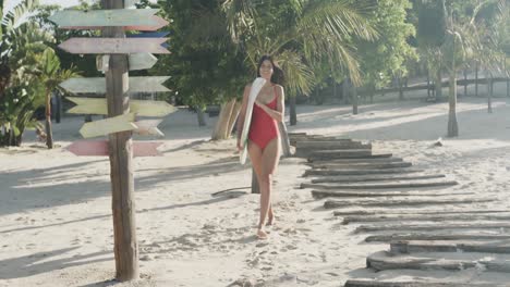 Feliz-Mujer-Hispana-En-Traje-De-Baño-Rojo-Llevando-Tabla-De-Surf-Caminando-En-La-Playa-Soleada,-Cámara-Lenta