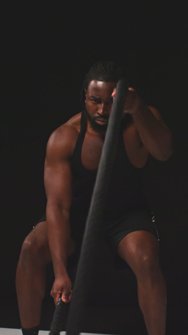 검은색 배경에 촬영된 전투 로프를 사용하여 피트니스 베스트를 입고 체육 훈련을 하는 남성 운동 선수의 수직 비디오 스튜디오