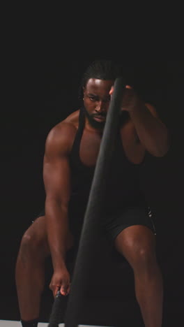 vertical video studio shot of male athlete gym training in fitness vest using battle ropes shot against black background