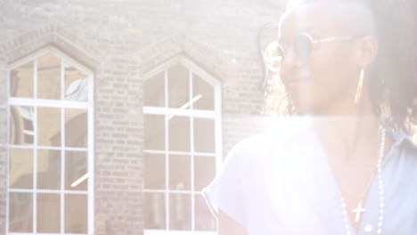 joven negra de moda en vestido azul y gafas de sol redondas caminando por la calle más allá de la cámara, retroiluminada