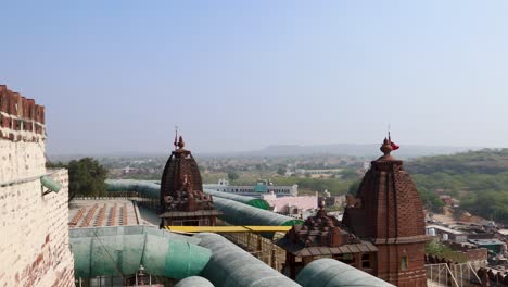 Antiguo-Templo-Hindú-Con-Una-Bandera-Sagrada-Ondeando-Y-Un-Fondo-De-Ciudad-Brumosa-Desde-La-Mañana-El-Video-Se-Toma-En-El-Templo-Osiyan-Sacchiyay-Mata,-Jodhpur,-Rajasthan,-India