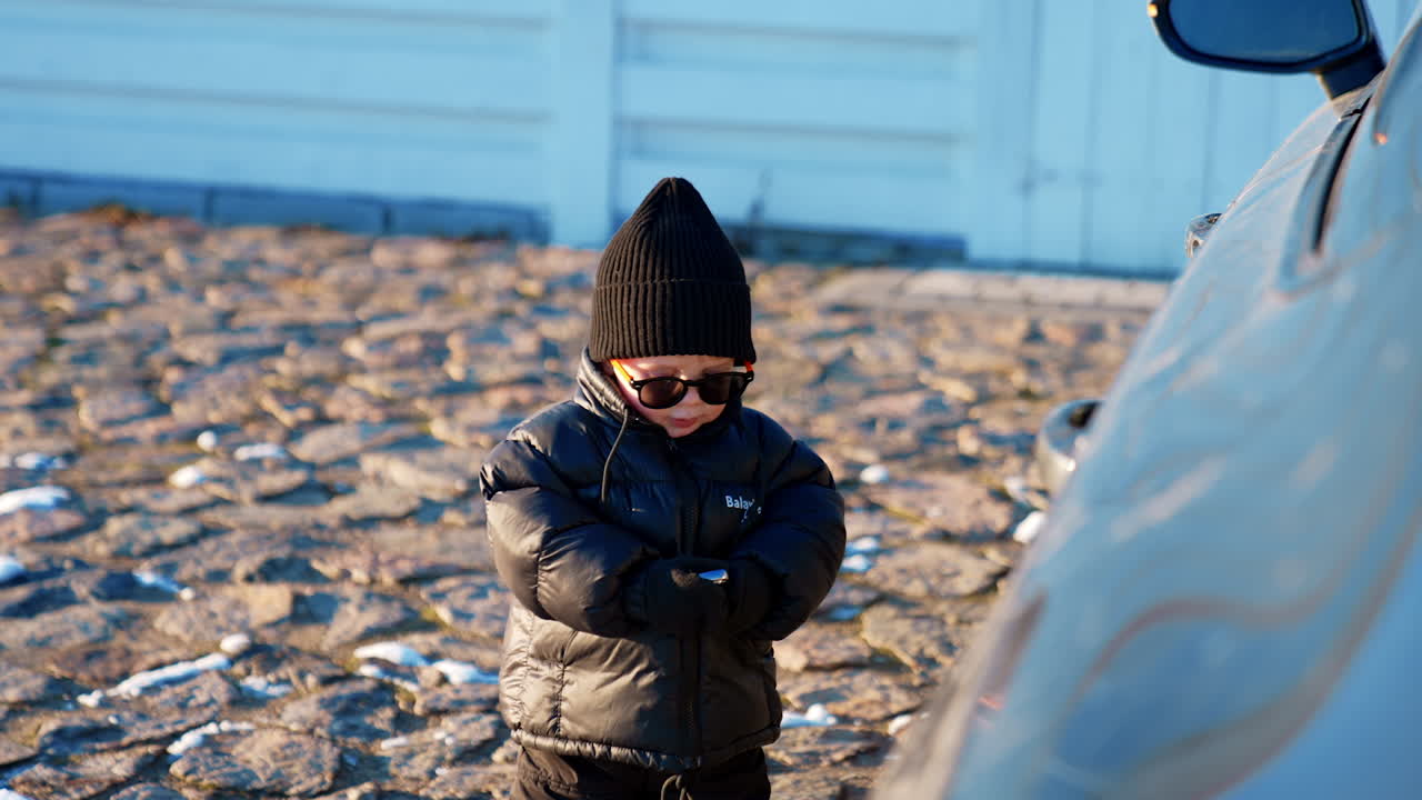 Fashionable little toddler wearing black cap, sunglasses and blazer stands at the car. Cute kid plays with car keys.